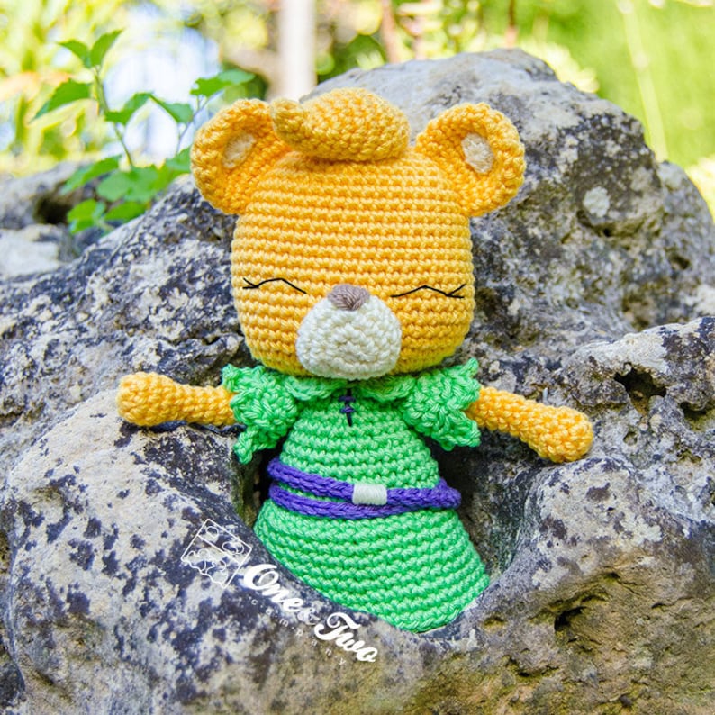 May include: A crocheted stuffed animal bear with a yellow head, green dress, and purple belt. The bear has a white face with black eyes and a closed mouth.