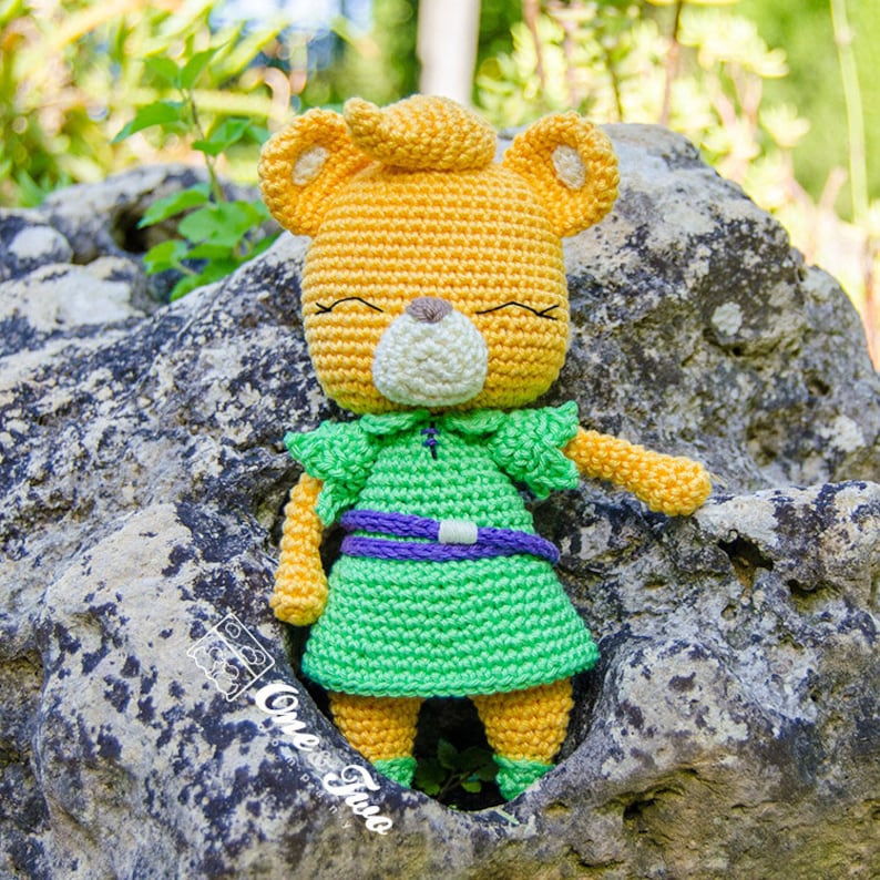 May include: A crocheted stuffed animal in the shape of a bear wearing a green dress with a purple belt and a white collar. The bear has a yellow body, white face, and closed eyes.