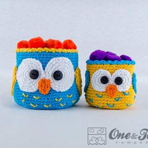 May include: Two handmade crochet owl-shaped containers. One is blue with orange and yellow accents, the other is yellow with blue and orange accents. Both have large white eyes, black button pupils, and orange beaks. The containers are filled with colorful pom-poms.