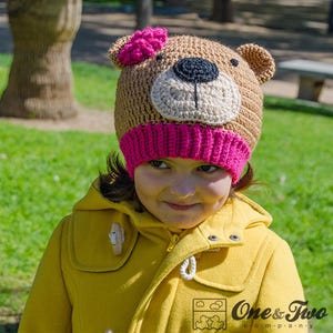 May include: A brown and pink crocheted bear hat with a pink flower on top. The hat is being worn by a child wearing a yellow jacket.