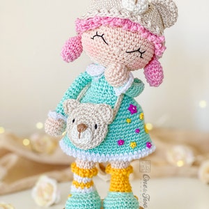 Könnte beinhalten: Eine gehäkelte Puppe, die ein hellblaues Kleid mit weißem Saum und einem braunen Bären-Applikationen trägt. Die Puppe trägt eine rosa-weiße Häkelmütze mit Bärenohren und gelbe und weiße gestreifte Socken. Die Puppe steht auf einem Korksockel mit dem Wort "Joy" darauf.