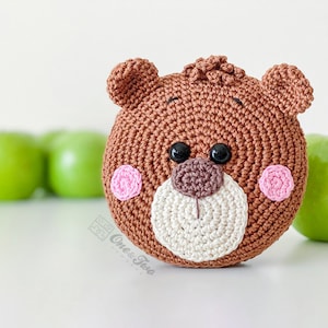 May include: A round, brown crocheted bear face with two small ears, a textured tuft of hair on top, black plastic eyes, a dark brown nose, a cream-coloured muzzle, and two pink circular cheeks. The text One & Two Company is printed on the lower left.
