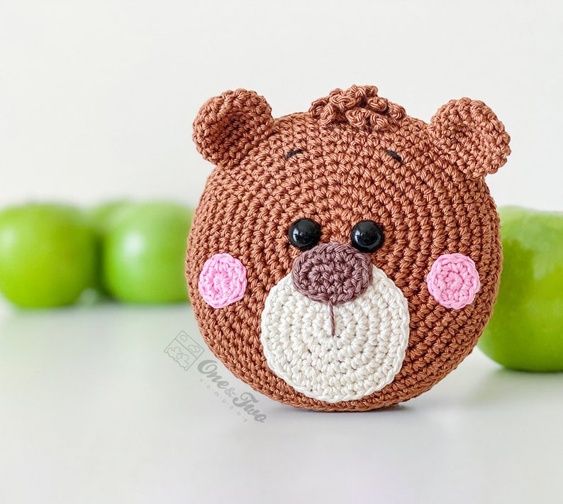 May include: A round, brown crocheted bear face with two small ears, a textured tuft of hair on top, black plastic eyes, a dark brown nose, a cream-coloured muzzle, and two pink circular cheeks. The text One & Two Company is printed on the lower left.
