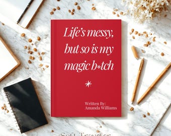 Könnte beinhalten: Ein rotes Buch mit dem Text "Life's messy, but so is my magic b*tch" und dem Namen der Autorin, Amanda Williams. Das Buch ist umgeben von Stiften, Notizbüchern und dekorativen Elementen auf einer Marmoroberfläche.