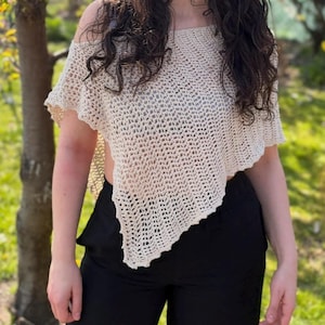 May include: A beige crochet top featuring an open-knit pattern with an asymmetrical hemline and short sleeves. The garment is worn with solid black trousers. The top has a loose, airy construction with a textured, repeating stitch design throughout.