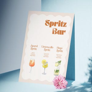 以下が含まれることがあります： オレンジ色の文字で「Spritz Bar」と書かれたメニューボード。アペロールスプリッツ、リモンチェッロスプリッツ、ヒューゴスプリッツの3種類のスプリッツカクテルのイラストが描かれています。ピンクの花が前景にあります。