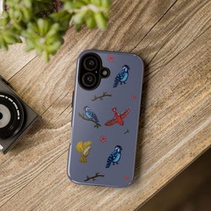 Op de afbeelding: Een leiblauwe smartphonehoes met een herhalend patroon van kleine vogels in blauw, geel en rood, afgewisseld met bruine boomtakken en kleine roze bloemetjes. De hoes heeft een ronde uitsparing voor de camera in de linkerbovenhoek.