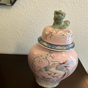 May include: A pink ceramic jar with a lid, featuring a detailed painting of birds and floral patterns. The lid is topped with a green, stylized lion figure. The jar has a blue and white decorative band around the lid.