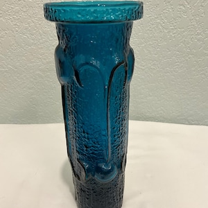 May include: A tall, textured teal glass vase. The vase has a cylindrical shape with a wide, textured rim. The surface of the vase has a rough, organic texture with rounded, decorative elements. The color is a deep, saturated teal.