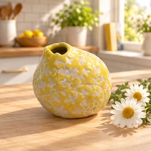 May include: A round, yellow ceramic vase featuring a textured, white floral pattern across its surface. The vase has a small, off-center opening at the top and a glossy finish, positioned on a wooden surface next to a small cluster of white daisies.