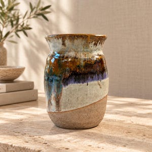 May include: A ceramic vase with a wide, flared rim and a rounded body. The upper portion features a glossy, multi-colored glaze in shades of deep brown, blue, and cream, while the lower section has a textured, unglazed, light beige clay finish.