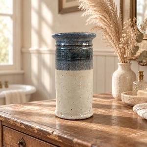 May include: A cylindrical ceramic vase featuring a two-tone glaze with a deep blue upper section and a speckled off-white lower section. The vase has a flared rim and a smooth, slightly textured finish, standing upright as a decorative vessel.