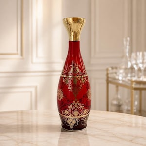 May include: A tall, slender glass vase featuring a deep red body adorned with intricate gold filigree patterns. The vase has a flared, polished gold-colored neck and a rounded base, showcasing a decorative design that wraps around the entire exterior.