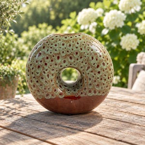 May include: A circular ceramic vase with a central opening, featuring a speckled light green and brown glaze on the upper portion and a solid dark reddish-brown glaze on the bottom half, creating a distinct contrast in texture and color.