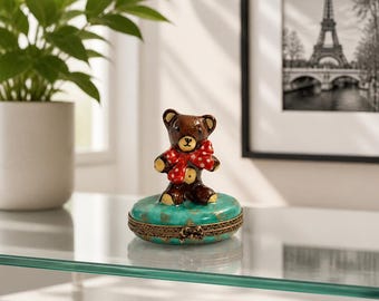 May include: A small, round, turquoise trinket box with a gold-toned metal rim and a hinged lid. A brown ceramic teddy bear figurine with a red and white polka-dot bow is attached to the top of the lid. The box features a distressed gold finish.