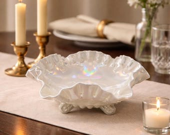 May include: A white iridescent glass bowl with a ruffled, scalloped edge and three small feet. The bowl features a glossy, pearlescent finish that reflects light with subtle rainbow hues, creating a shimmering effect across its curved surface.
