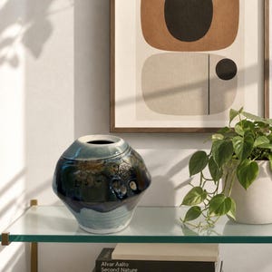 May include: A round ceramic vase with a glossy finish featuring a gradient of deep blue, teal, and white tones. The vase has a wide, bulbous body that tapers toward a narrow opening at the top, showcasing a textured, mottled surface pattern.