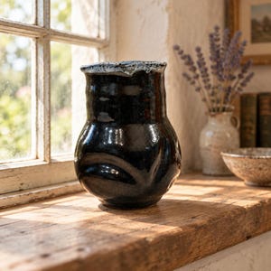 May include: A handmade ceramic vase with a glossy black finish and an irregular, organic shape. The rim features a contrasting white and blue speckled glaze, creating a distinct border against the dark, reflective body of the vessel.