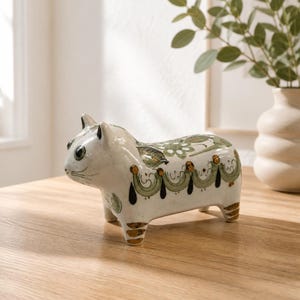May include: A white ceramic cat figurine featuring hand-painted green floral patterns and decorative gold-colored accents along the back and legs. The cat has large dark eyes, pointed ears, and a smooth, glossy finish on its four-legged body.