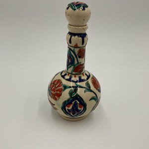 May include: A ceramic bottle with a bulbous base and a long neck, adorned with red, blue, and green floral patterns. The bottle has a cream-colored background and a matching stopper. The design is reminiscent of traditional pottery.