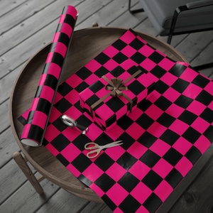Peut inclure: Rouleau de papier cadeau à carreaux roses et noirs, avec une boîte cadeau assortie et une feuille de papier. Des ciseaux dorés et un distributeur de ruban adhésif argenté sont également sur la table. La boîte cadeau est attachée avec un ruban marron.