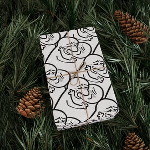 May include: A wrapped gift with white paper featuring a repeating black line drawing of a sad face. The gift is tied with twine and rests on a bed of green pine needles with several brown pine cones.