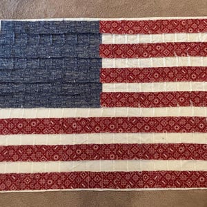 May include: A handmade American flag quilt. The upper left corner features a blue field with a textured pattern. The flag has red and white stripes, with a red paisley pattern on the red stripes. The quilt is rectangular.