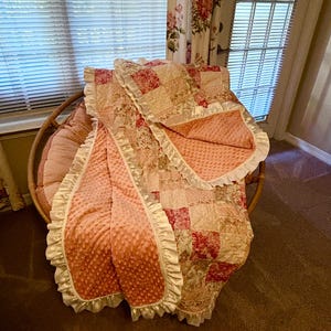 May include: A patchwork quilt and a matching blanket are draped over a wicker chair. The quilt features a mix of floral and solid fabric squares in shades of pink, red, and cream. The blanket has a textured, dimpled surface and a ruffled edge.