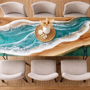 May include: A wooden dining table with a turquoise resin river design, resembling ocean waves. The table is surrounded by six light grey chairs and a woven rug. A decorative tray with candles and dried plants sits on the table.