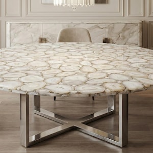 May include: A large, round table with a unique surface made of white and beige agate slices. The table has a polished, metallic, X-shaped base. The table is a statement piece for a dining room.