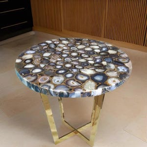 May include: A round side table with a top made of polished agate slices in shades of blue, grey, and white. The table has a gold-coloured metal base with an X-shaped design. The table is approximately 50 cm in diameter.