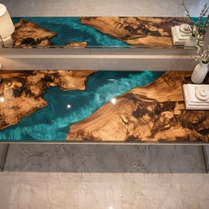 Op de afbeelding: Een moderne consoletafel met een uniek ontwerp. Het tafelblad heeft een combinatie van natuurlijk hout en turquoise hars, waardoor een rivierachtig effect ontstaat. De tafel wordt ondersteund door een slank, zilverkleurig metalen frame. De tafel is ongeveer 152 cm lang.