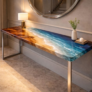Op de afbeelding: Een rechthoekige consoletafel met een epoxyhars blad met strandthema. De bovenkant gaat over van een zanderig bruin naar een levendig blauw, dat de oceaangolven nabootst. De tafel heeft een gepolijst metalen frame en is versierd met een kleine lamp en vaas.