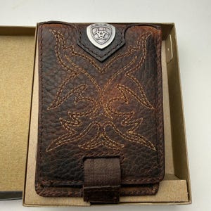 May include: Brown leather wallet with decorative stitching and a metal emblem. The wallet features a stitched design and a brown fabric strap. The wallet is in a cardboard box.