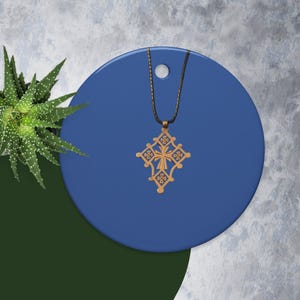 May include: A gold-colored pendant necklace featuring an ornate cross design. The cross is suspended from a black cord and rests on a blue circular surface. A green plant is visible in the background.