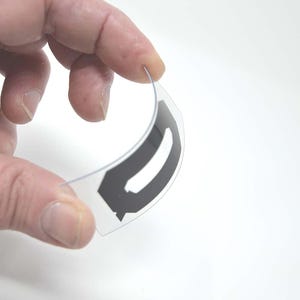 Puede incluir: Un componente de pl&aacute;stico transparente y curvado con un dise&ntilde;o geom&eacute;trico negro. El objeto se sostiene entre los dedos sobre un fondo blanco, mostrando su forma y el contraste entre los elementos transparentes y negros.
