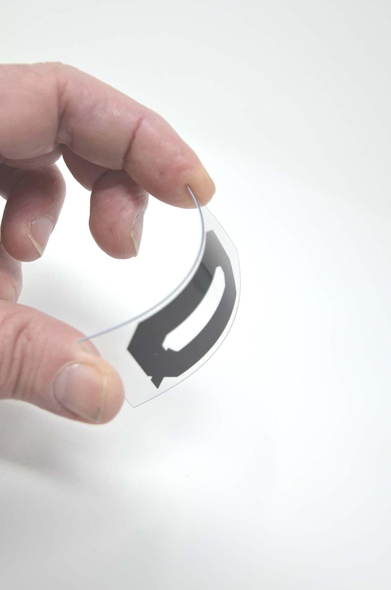 Puede incluir: Un componente de pl&aacute;stico transparente y curvado con un dise&ntilde;o geom&eacute;trico negro. El objeto se sostiene entre los dedos sobre un fondo blanco, mostrando su forma y el contraste entre los elementos transparentes y negros.