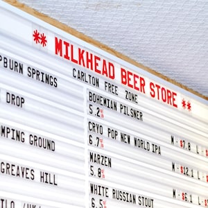 Può includere: Un'insegna luminosa bianca con il testo rosso "MILKHEAD BEER STORE". L'insegna elenca varie birre, tra cui Bohemian Pilsner, Cryo Pop New World IPA, Marzen e White Russian Stout, con le loro percentuali alcoliche.