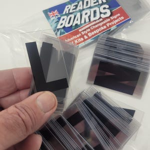 Puede incluir: Letras de se&ntilde;alizaci&oacute;n negras en fundas de pl&aacute;stico transparente. La imagen muestra varios paquetes de letras, algunos de ellos sostenidos en una mano. El embalaje presenta el texto "READER BOARDS" y una bandera brit&aacute;nica.