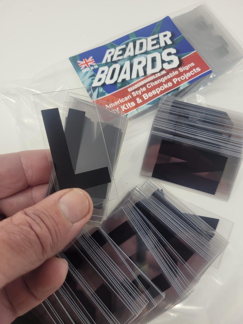 Puede incluir: Letras de se&ntilde;alizaci&oacute;n negras en fundas de pl&aacute;stico transparente. La imagen muestra varios paquetes de letras, algunos de ellos sostenidos en una mano. El embalaje presenta el texto "READER BOARDS" y una bandera brit&aacute;nica.