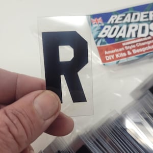 Puede incluir: Una pegatina negra con la letra "R" sobre un fondo transparente. La pegatina se sostiene frente a una pila de pegatinas similares. El logotipo de Reader Boards es visible en el fondo.