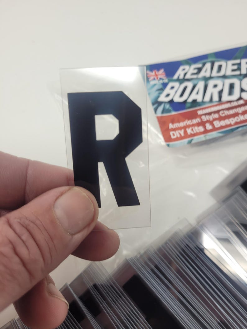 Puede incluir: Una pegatina negra con la letra "R" sobre un fondo transparente. La pegatina se sostiene frente a una pila de pegatinas similares. El logotipo de Reader Boards es visible en el fondo.