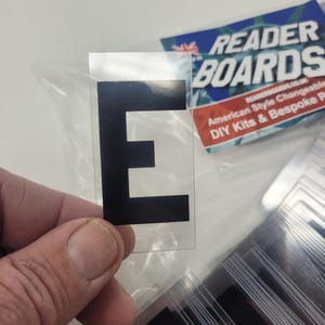Puede incluir: Una pegatina transparente con la letra negra &laquo;E&raquo; se sostiene en la mano. La pegatina forma parte de un conjunto, con otras pegatinas similares visibles en el fondo. Tambi&eacute;n se ve un letrero de Reader Boards.