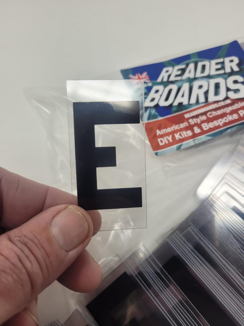 Puede incluir: Una pegatina transparente con la letra negra &laquo;E&raquo; se sostiene en la mano. La pegatina forma parte de un conjunto, con otras pegatinas similares visibles en el fondo. Tambi&eacute;n se ve un letrero de Reader Boards.