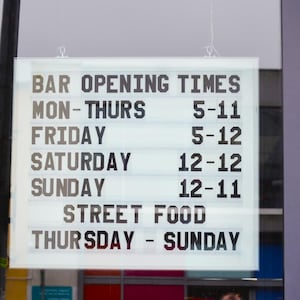 Puede incluir: Un letrero luminoso muestra los horarios de apertura de un bar: Lun-Jue 5-11, Vie 5-12, S&aacute;b 12-12, Dom 12-11. Comida callejera disponible de jueves a domingo. Texto negro sobre fondo blanco.