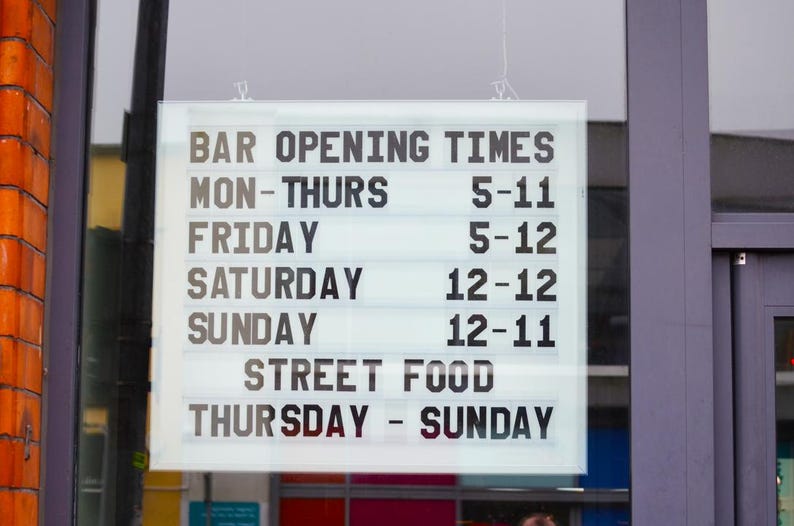 Puede incluir: Un letrero luminoso muestra los horarios de apertura de un bar: Lun-Jue 5-11, Vie 5-12, S&aacute;b 12-12, Dom 12-11. Comida callejera disponible de jueves a domingo. Texto negro sobre fondo blanco.