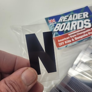 Puede incluir: Una letra "N" de pl&aacute;stico transparente con relleno negro, sostenida en una mano. La letra es parte de un conjunto de letras intercambiables. Un letrero azul y blanco en el fondo dice "READER BOARDS".
