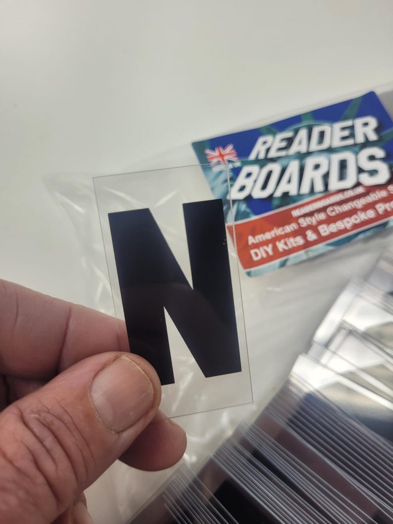 Puede incluir: Una letra "N" de pl&aacute;stico transparente con relleno negro, sostenida en una mano. La letra es parte de un conjunto de letras intercambiables. Un letrero azul y blanco en el fondo dice "READER BOARDS".