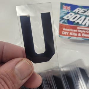 Puede incluir: Una pegatina negra con la letra "U" sobre un soporte transparente, sostenida en una mano. La pegatina forma parte de un conjunto, con otras letras visibles. Una etiqueta parcialmente visible dice "Ready Board".