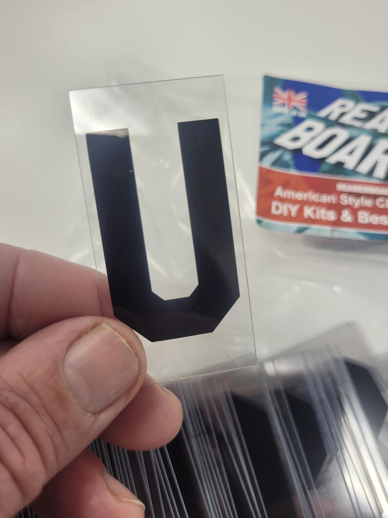 Puede incluir: Una pegatina negra con la letra "U" sobre un soporte transparente, sostenida en una mano. La pegatina forma parte de un conjunto, con otras letras visibles. Una etiqueta parcialmente visible dice "Ready Board".